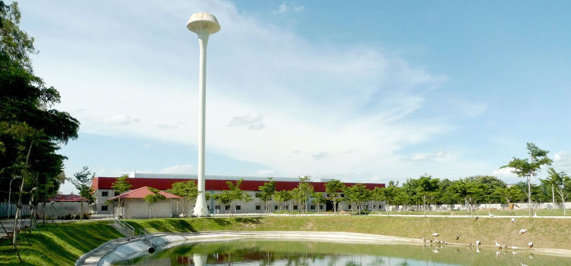 A wide outdoor scene showing a tall white tower beside a pond, with grassy slopes, trees, and a long building with a red roof in the background under a blue sky.