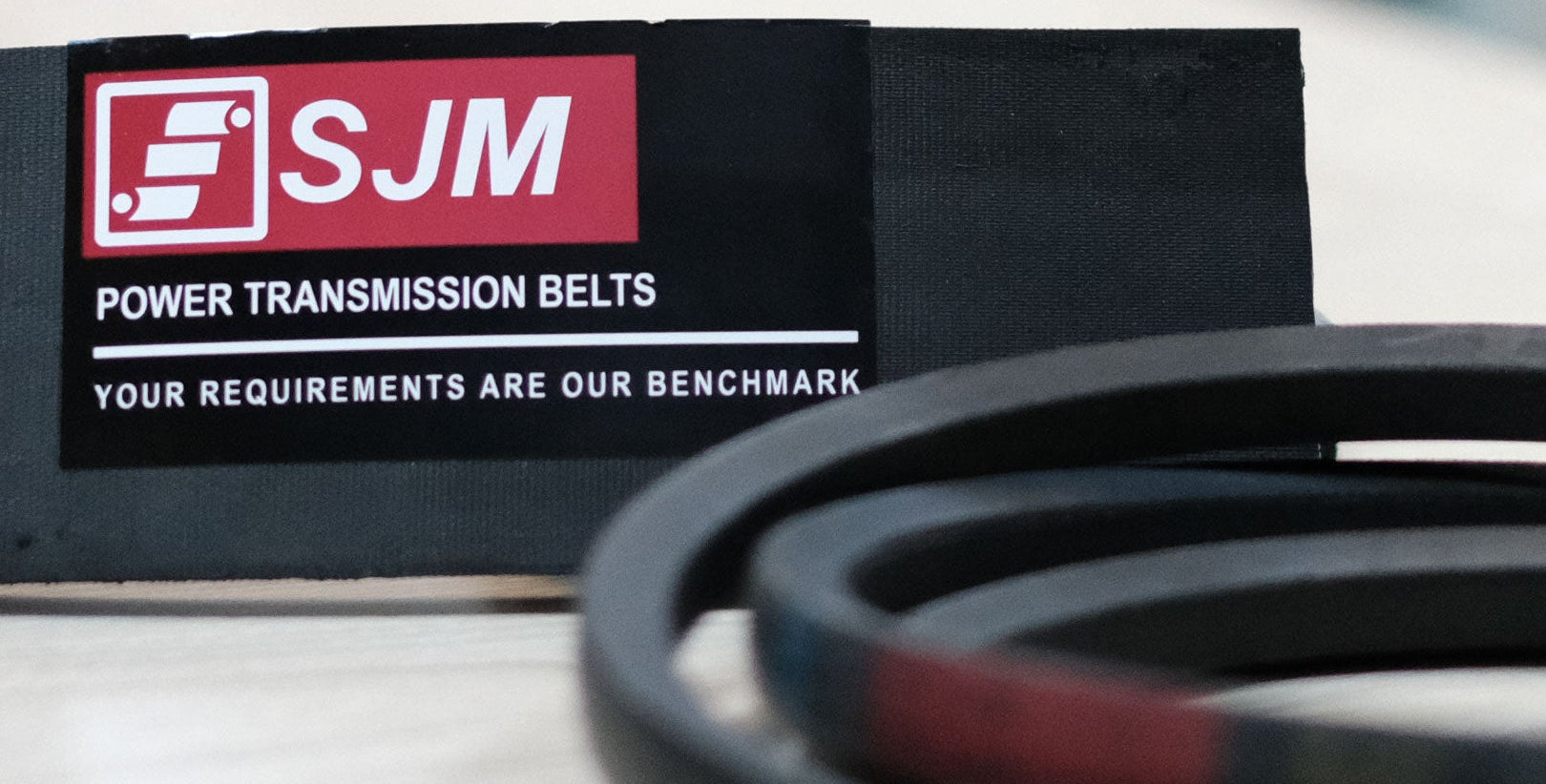 A close-up of a coiled black power transmission belt in the foreground with a black fabric label in the background featuring the red and white SJM logo and the text 'POWER TRANSMISSION BELTS' and 'YOUR REQUIREMENTS ARE OUR BENCHMARK'.